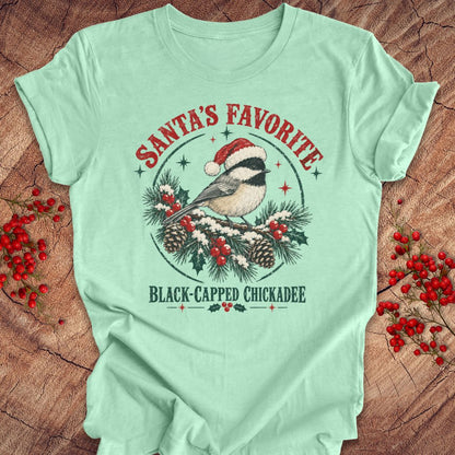 Heather mint green t-shirt with a Black-capped Chickadee in a Santa hat on a snowy pine bough with holly and red berries, 'Santa's Favorite' and 'Black-capped Chickadee' text, laid flat on a rustic wood background with red winter berries.