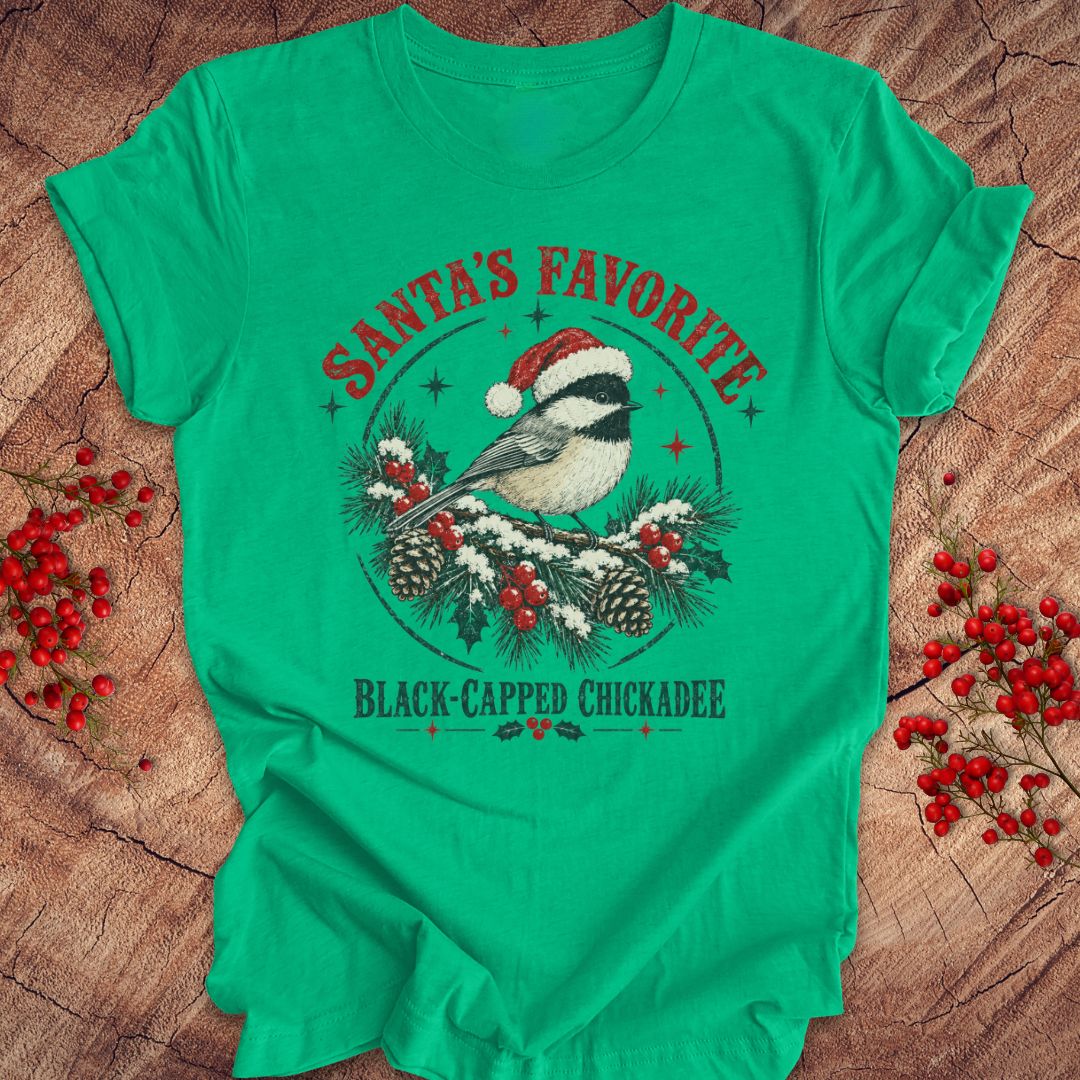 Heather kelly green t-shirt with a Black-capped Chickadee in a Santa hat on a snowy pine bough with holly and red berries, 'Santa's Favorite' and 'Black-capped Chickadee' text, laid flat on a rustic wood background with red winter berries.