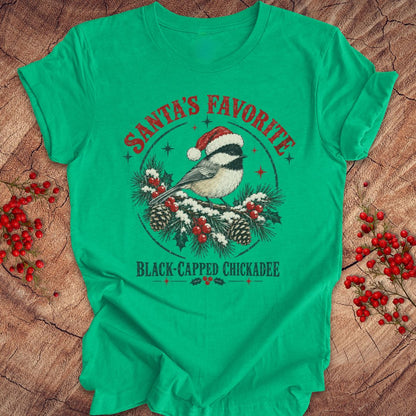 Heather kelly green t-shirt with a Black-capped Chickadee in a Santa hat on a snowy pine bough with holly and red berries, 'Santa's Favorite' and 'Black-capped Chickadee' text, laid flat on a rustic wood background with red winter berries.