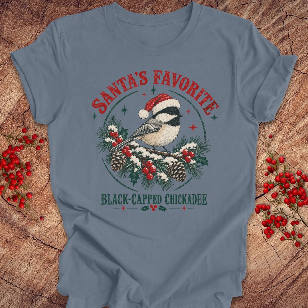 Steel blue t-shirt with a Black-capped Chickadee in a Santa hat on a snowy pine bough with holly and red berries, 'Santa's Favorite' and 'Black-capped Chickadee' text, laid flat on a rustic wood background with red winter berries.