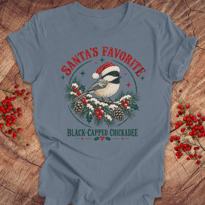 Steel blue t-shirt with a Black-capped Chickadee in a Santa hat on a snowy pine bough with holly and red berries, 'Santa's Favorite' and 'Black-capped Chickadee' text, laid flat on a rustic wood background with red winter berries.