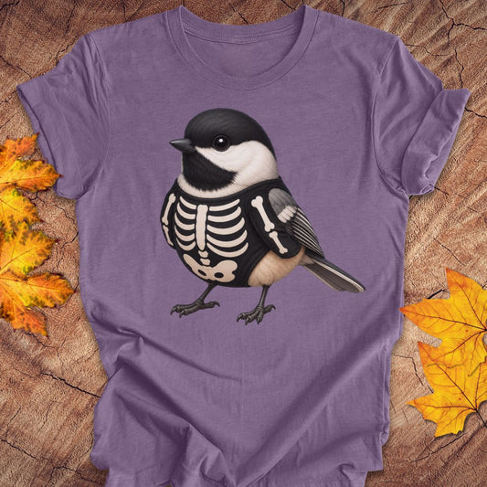 A purple short-sleeve crewneck t-shirt featuring a graphic of a chickadee bird wearing a black-and-white skeleton outfit, laid flat on a rustic wood background with autumn leaves.