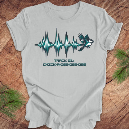 Silver t-shirt with a chickadee and sound wave design on a wooden surface