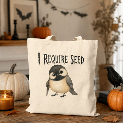 A cream canvas tote bag with a printed illustration of a bird and the text 'I REQUIRE SEED' sits on a wooden table in a Halloween-themed setting with pumpkins, a candle, and a black bird figurine.