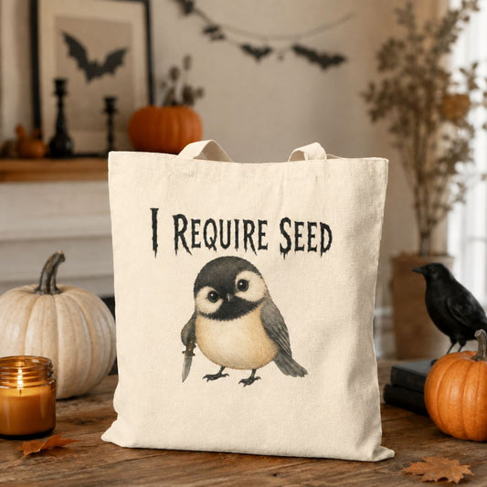 A cream canvas tote bag with a printed illustration of a bird and the text 'I REQUIRE SEED' sits on a wooden table in a Halloween-themed setting with pumpkins, a candle, and a black bird figurine.