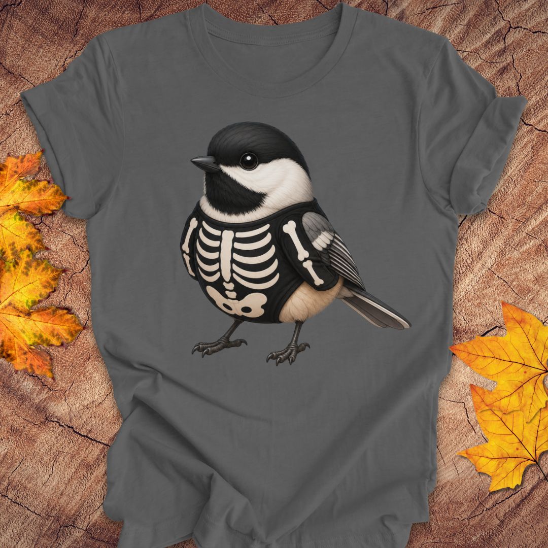 A dark gray t-shirt featuring a graphic of a chickadee bird wearing a black and white skeleton outfit, displayed on a rustic wood background with autumn leaves.