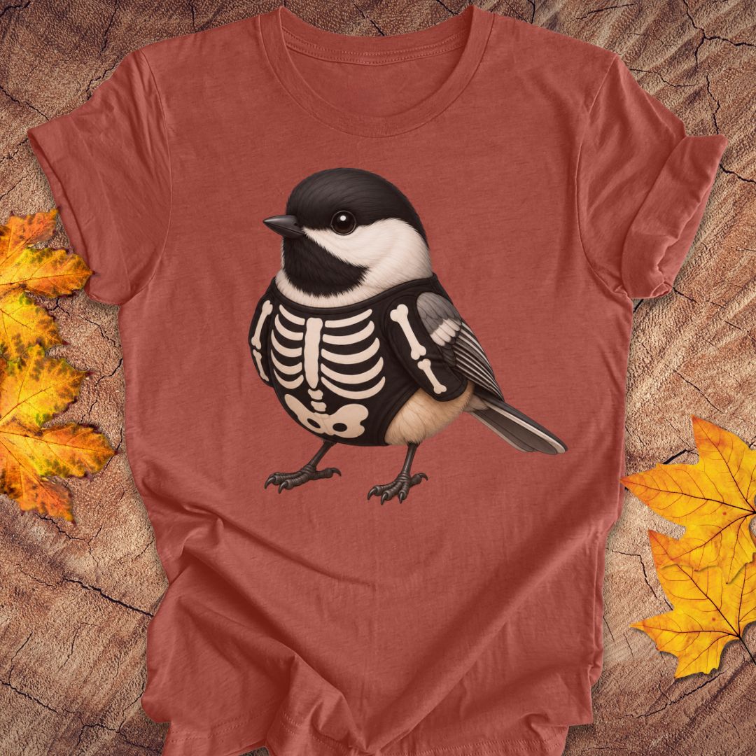 A terracotta-colored short-sleeve t-shirt featuring a graphic of a black and white chickadee bird with a skeleton print on its back, displayed on a rustic wood background with autumn leaves.
