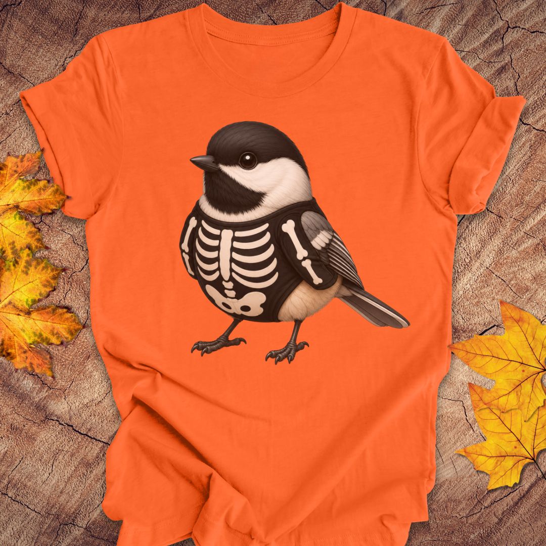 An orange short-sleeve T-shirt laid flat on a rustic wood surface with autumn leaves around it. The shirt features a large graphic of a black-and-white chickadee bird with a skeleton chest design.