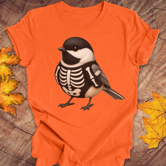 An orange short-sleeve T-shirt laid flat on a rustic wood surface with autumn leaves around it. The shirt features a large graphic of a black-and-white chickadee bird with a skeleton chest design.