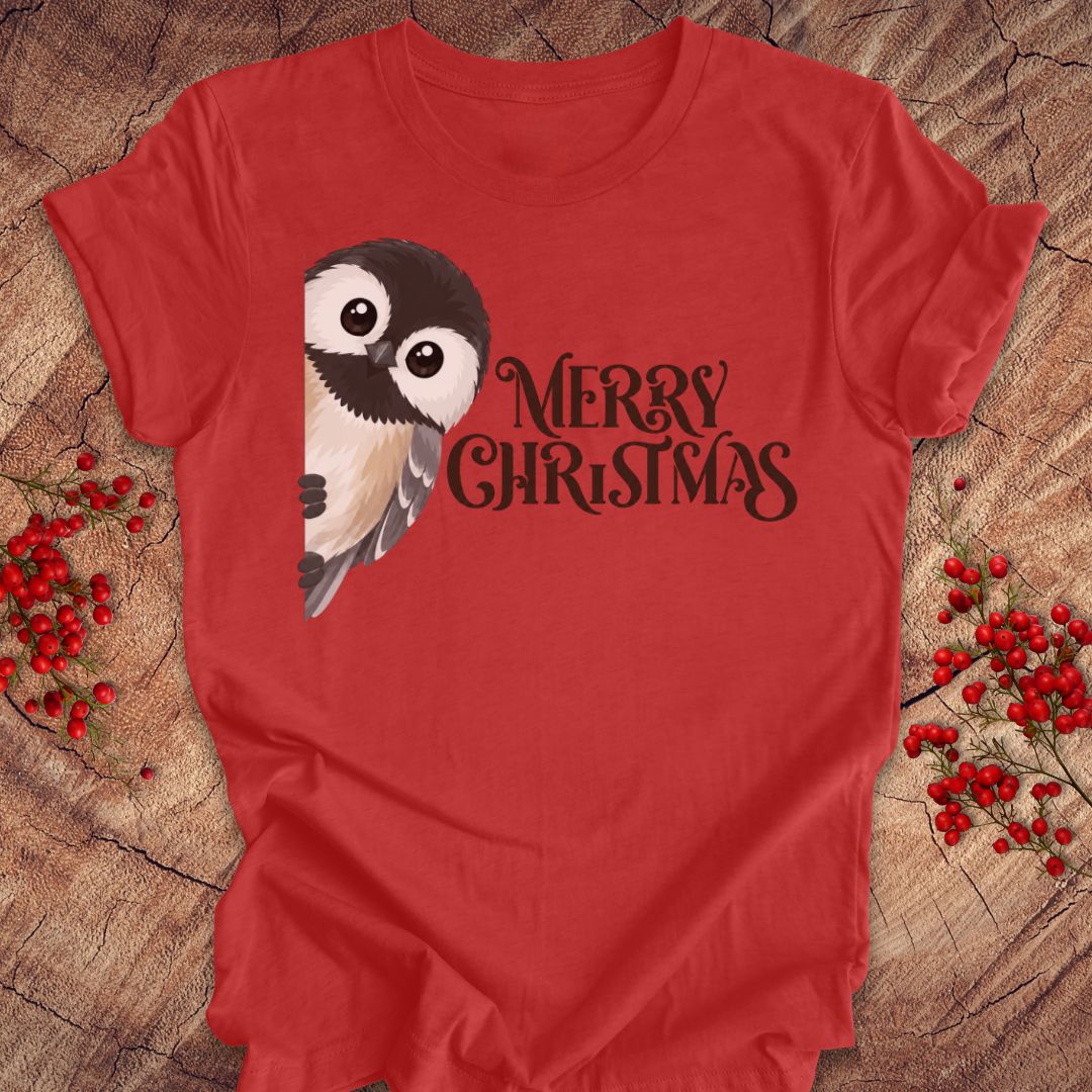 A red short-sleeve t-shirt laid flat on a rustic wood surface, featuring a black-and-white bird illustration and the words 'MERRY CHRISTMAS' in black script. Red berry sprigs are placed around the shirt as styling props.