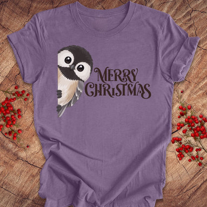 A purple short-sleeve t-shirt laid flat on a rustic wood background, decorated with red berries. The shirt features a graphic of a cute cartoon owl and the text 'MERRY CHRISTMAS' in dark brown.