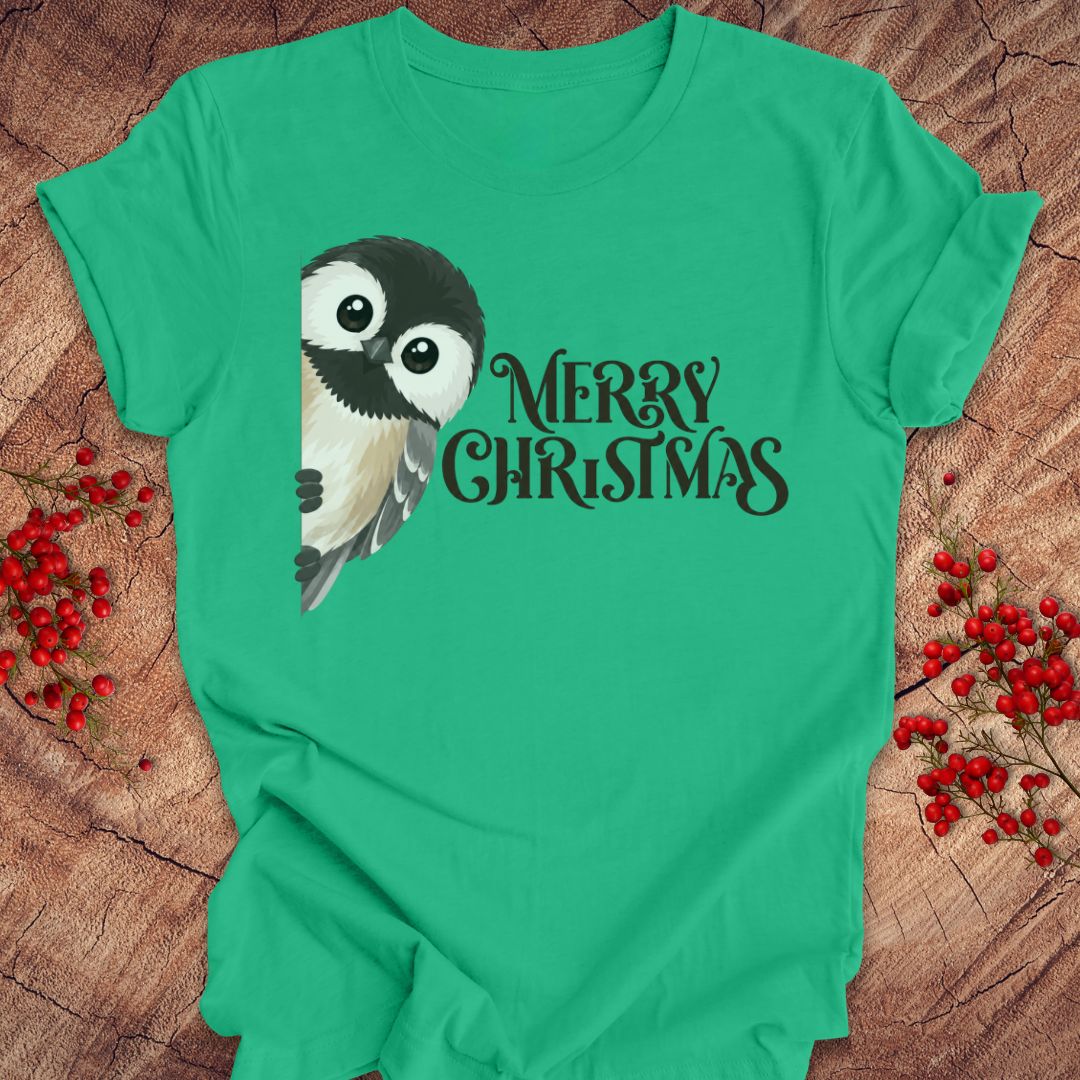 A bright green short-sleeve T-shirt laid flat on a rustic wood surface, featuring a black-and-white bird illustration and the words “MERRY CHRISTMAS” in black script. Red berry sprigs are placed around the shirt as styling props.