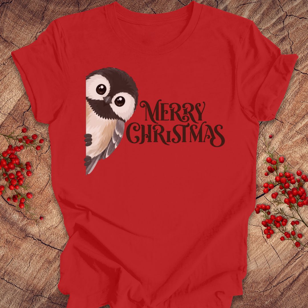 A red short-sleeve t-shirt laid flat on a rustic wood surface, featuring a black-and-white bird illustration and the words 'MERRY CHRISTMAS' in black script. Red berry sprigs are placed around the shirt as styling props.