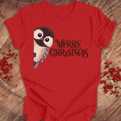 A red short-sleeve t-shirt laid flat on a rustic wood surface, featuring a black-and-white bird illustration and the words 'MERRY CHRISTMAS' in black script. Red berry sprigs are placed around the shirt as styling props.