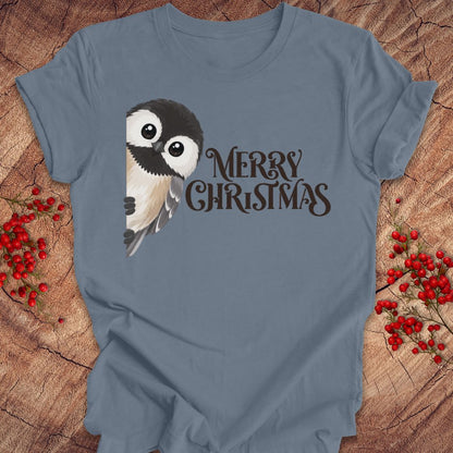 A slate blue short-sleeve t-shirt with a graphic of a cute black and white bird next to the text 'MERRY CHRISTMAS' in dark brown, laid flat on a rustic wood background with red berries.