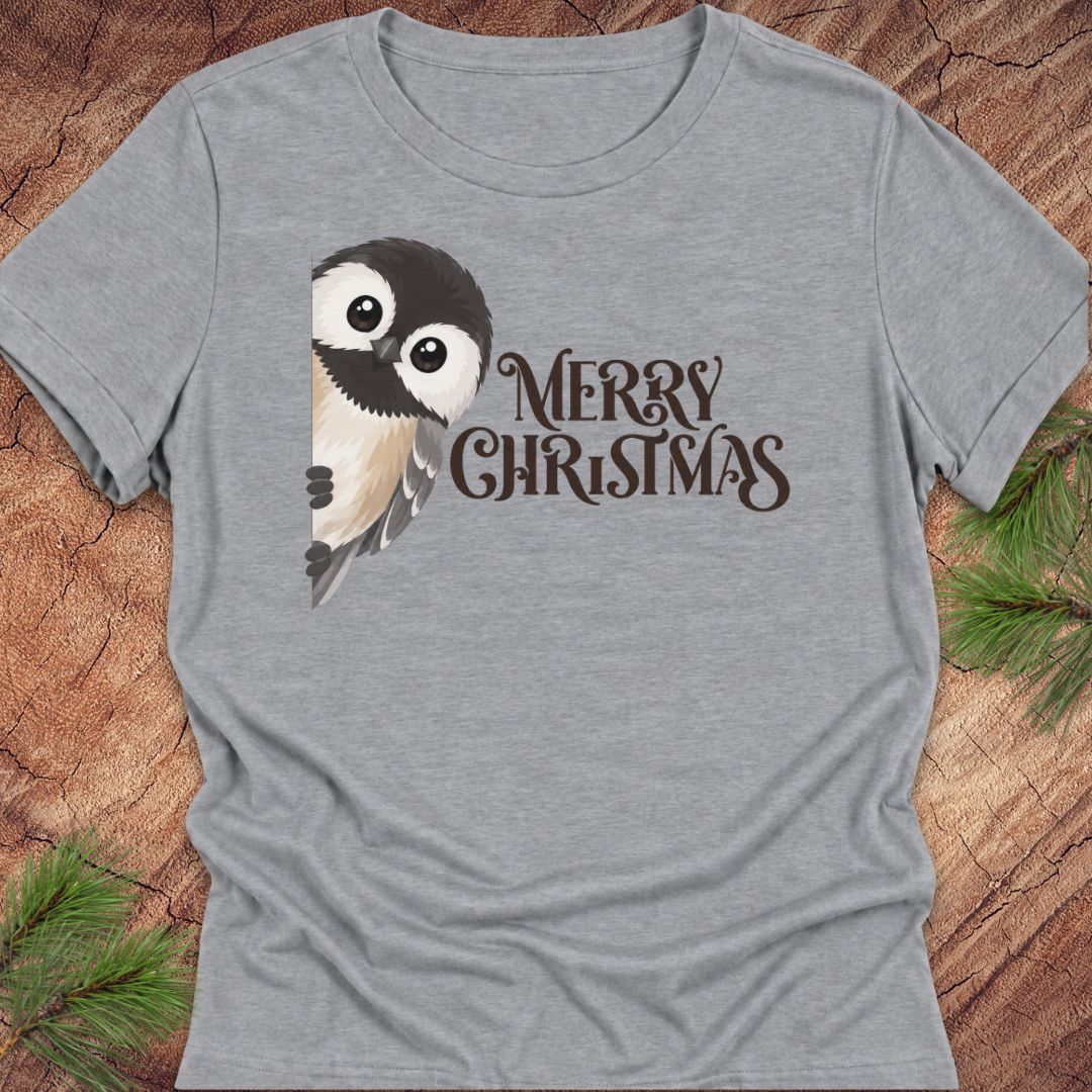 A heather gray short-sleeve t-shirt with a graphic of a cute bird owl and the text 'MERRY CHRISTMAS' in dark brown, laid flat on a rustic wood background with pine needles.