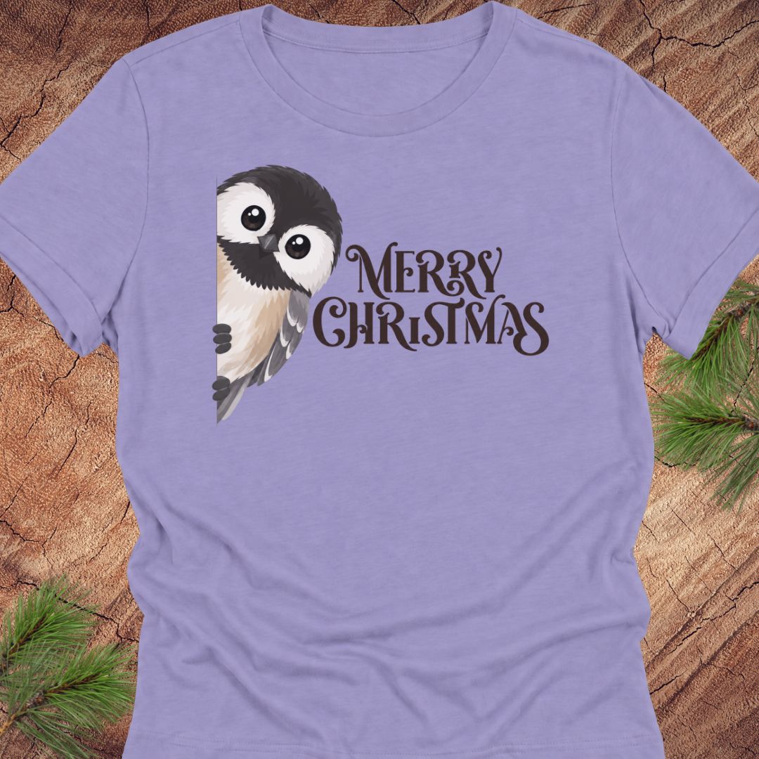 A lavender purple short-sleeve t-shirt featuring a cute cartoon chickadee and the text 'MERRY CHRISTMAS' in dark brown, laid flat on a rustic wood background with pine needles.