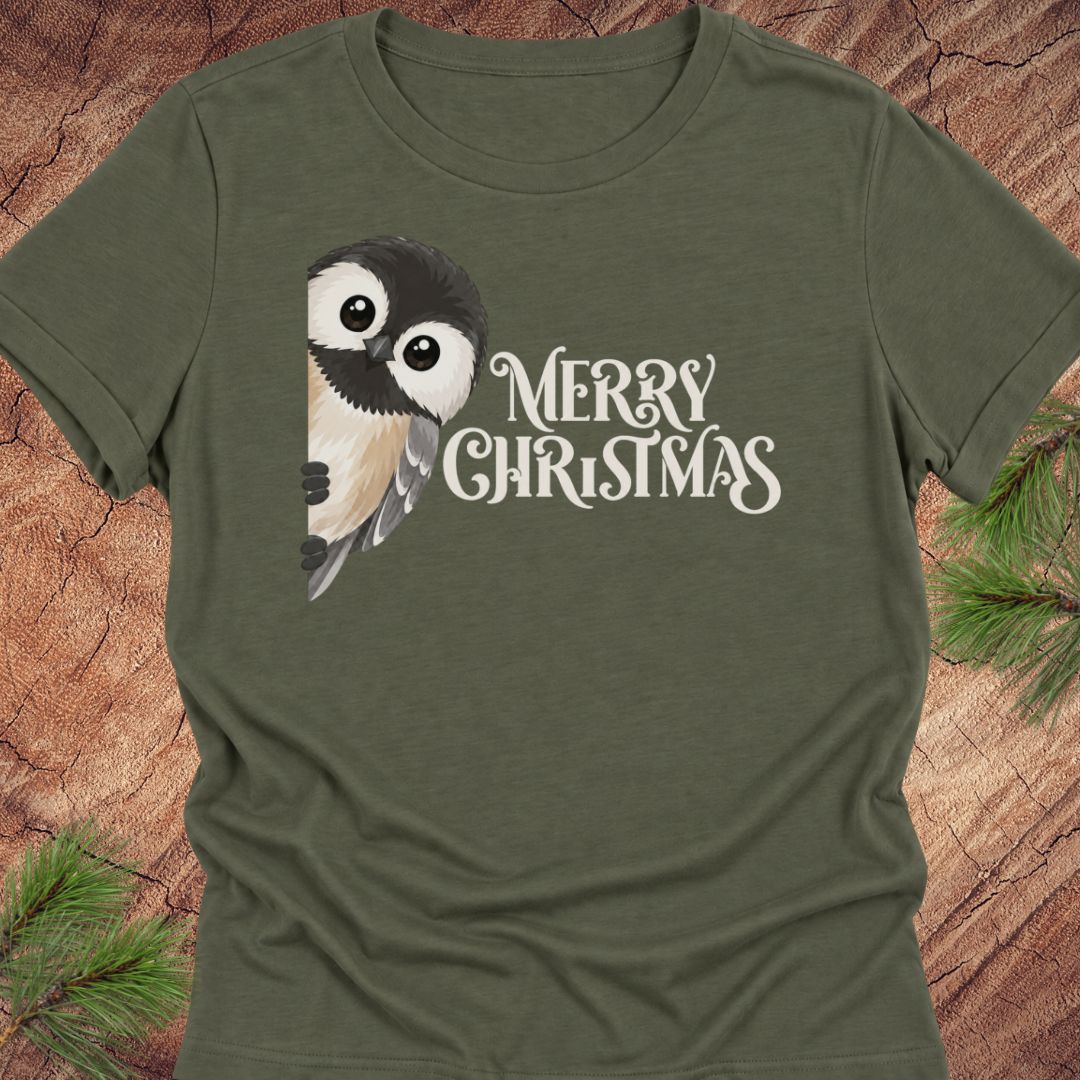 An olive green short-sleeve t-shirt featuring a cartoon chickadee and the text 'MERRY CHRISTMAS' in white, laid flat on a rustic wood background with pine needles in the corners.