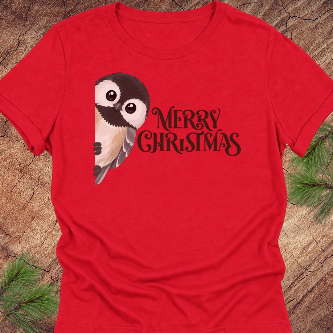 A red short-sleeve crewneck t-shirt featuring a cute cartoon owl graphic and the text 'MERRY CHRISTMAS' in white script, laid flat on a rustic wood background with pine needles in the corners.