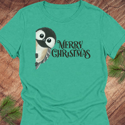 A teal green short-sleeve t-shirt featuring a cute cartoon owl and the text 'MERRY CHRISTMAS' in black, laid flat on a rustic wood background with pine needles.