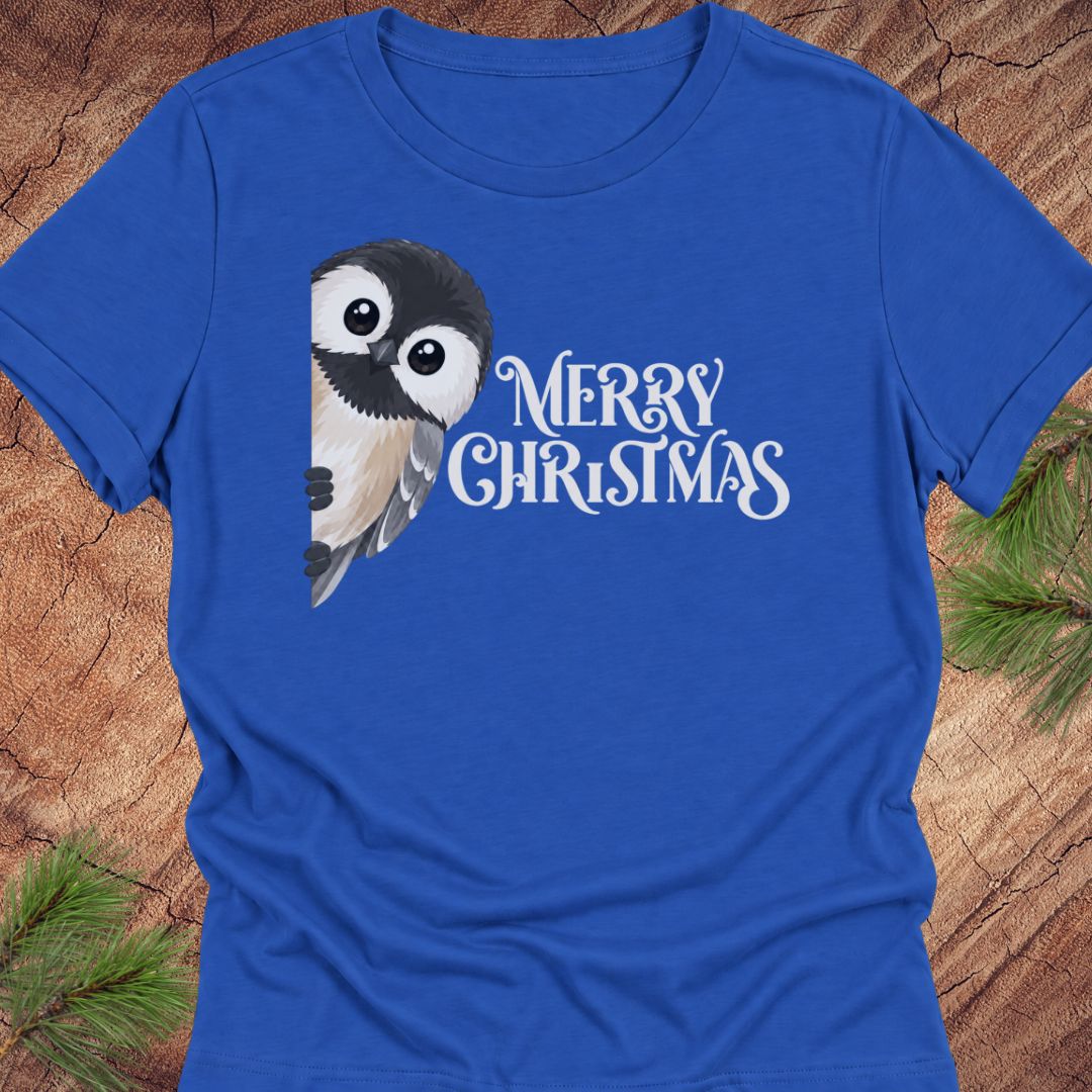 A royal blue short-sleeve crewneck t-shirt featuring a cute cartoon owl graphic and the text 'MERRY CHRISTMAS' in white script, laid flat on a rustic wood background with pine needles in the corners.