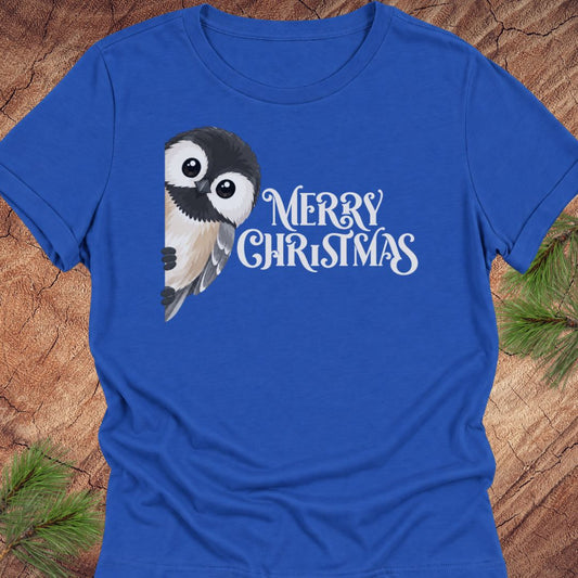 A royal blue short-sleeve crewneck t-shirt featuring a cute cartoon owl graphic and the text 'MERRY CHRISTMAS' in white script, laid flat on a rustic wood background with pine needles in the corners.