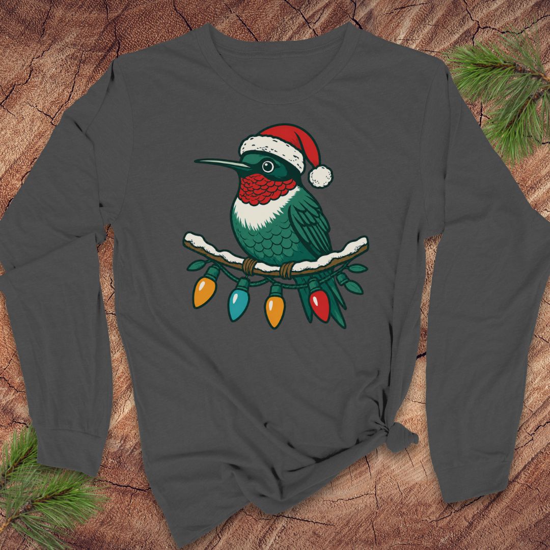 A dark gray long-sleeve t-shirt with a graphic of a Christmas hummingbird wearing a Santa hat, displayed on a rustic wood background with pine branches.