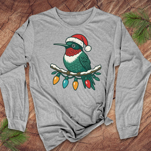 A heather gray long-sleeve t-shirt with a graphic of a hummingbird wearing a Santa hat and perched on a branch with Christmas lights, laid flat on a rustic wood background with pine needles.