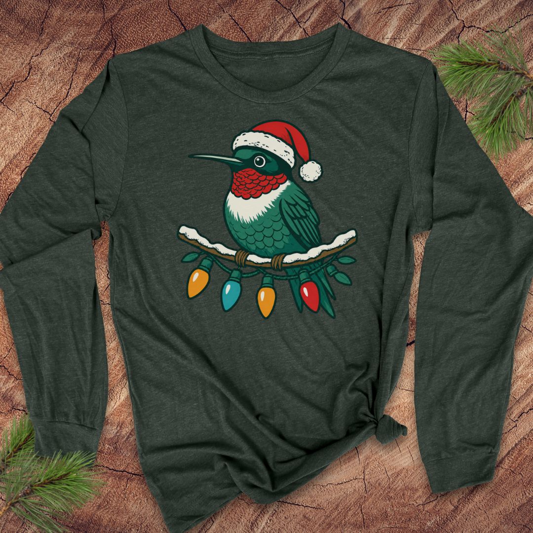 A dark forest green long-sleeve t-shirt featuring a graphic of a hummingbird wearing a red Santa hat and perched on Christmas lights, laid flat on a rustic wood background with pine branches.