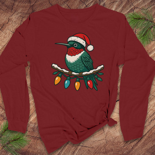 A dark red long-sleeve t-shirt with a graphic of a green hummingbird wearing a red Santa hat and perched on Christmas lights, laid flat on a rustic wood background with pine branches.