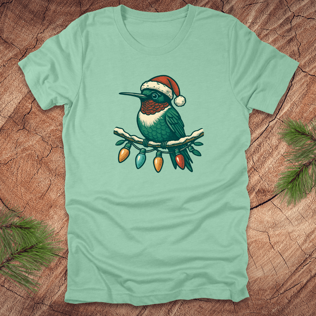 Mint green t-shirt of a hummingbird in a Santa hat sitting on a winter branch with christmas lights on a wooden background.