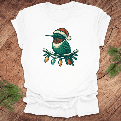 White t-shirt of a hummingbird in a Santa hat sitting on a winter branch with christmas lights on a wooden background.