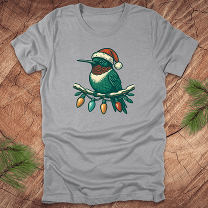Grey t-shirt of a hummingbird in a Santa hat sitting on a winter branch with christmas lights on a wooden background.