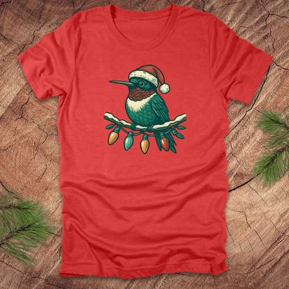 Red t-shirt of a hummingbird in a Santa hat sitting on a winter branch with christmas lights on a wooden background.