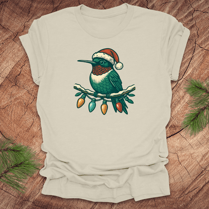 Beige t-shirt of a hummingbird in a Santa hat sitting on a winter branch with christmas lights on a wooden background.