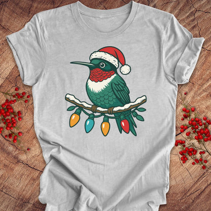 A light gray short-sleeve t-shirt laid flat on a rustic wood background, featuring a graphic of a green hummingbird wearing a red Santa hat perched on a branch with multicolored Christmas lights.