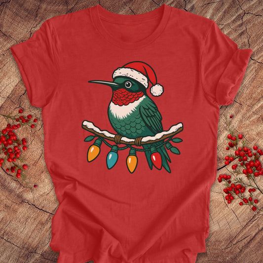A red short-sleeve T-shirt laid flat on a rustic wood surface, featuring a colorful graphic of a green hummingbird wearing a red Santa hat perched on a branch with hanging Christmas lights. Small red berry sprigs are placed around the shirt.