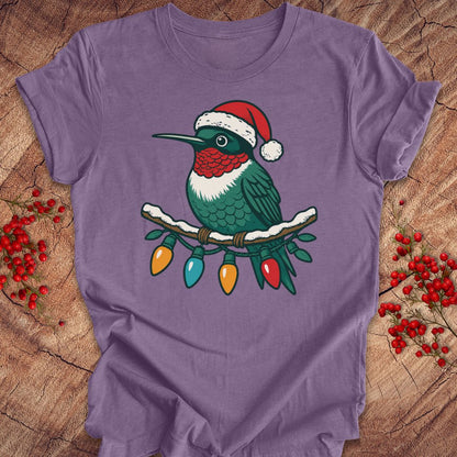 A purple short-sleeve t-shirt laid flat on a rustic wood surface, featuring a graphic of a green hummingbird wearing a red Santa hat perched on a branch with colorful Christmas lights. Red berry sprigs are placed on both sides of the shirt.