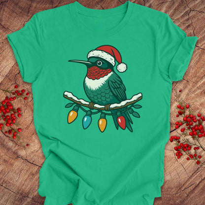 A green short-sleeve T-shirt laid flat on a rustic wood surface, featuring a graphic of a hummingbird wearing a red Santa hat perched on a branch with multicolored Christmas lights. Red berry sprigs are placed around the shirt.