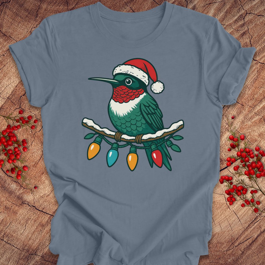 A slate blue short-sleeve t-shirt laid flat on a rustic wood background, featuring a graphic of a green hummingbird wearing a red Santa hat with Christmas lights hanging from its beak.
