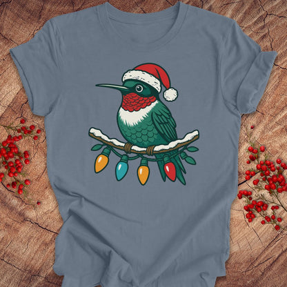 A slate blue short-sleeve t-shirt laid flat on a rustic wood background, featuring a graphic of a green hummingbird wearing a red Santa hat with Christmas lights hanging from its beak.