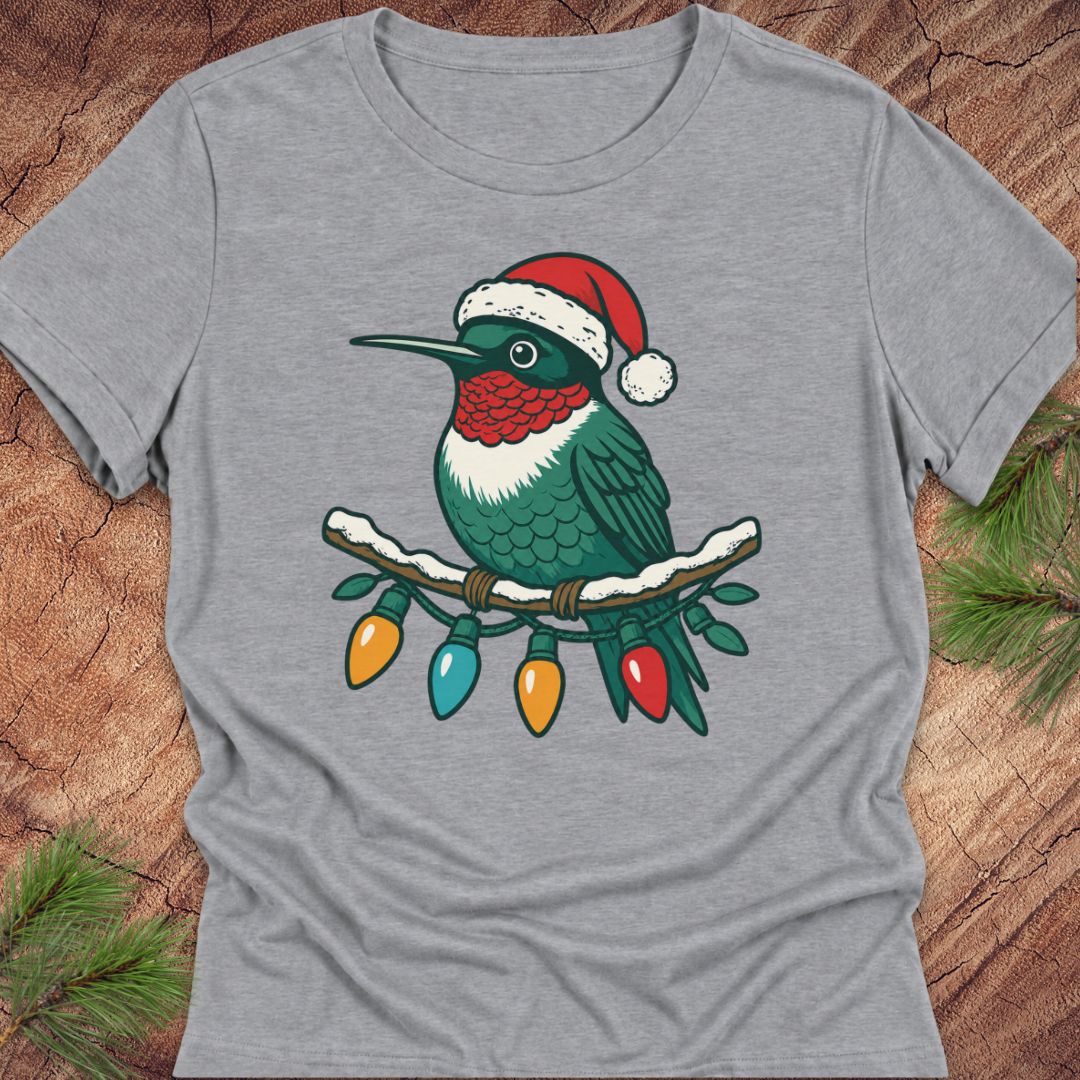 A heather gray short-sleeve t-shirt laid flat on a rustic wood background with pine branches in the corners. The shirt features a graphic of a green hummingbird wearing a red Santa hat, perched on a snowy branch with hanging multicolored Christmas lights.