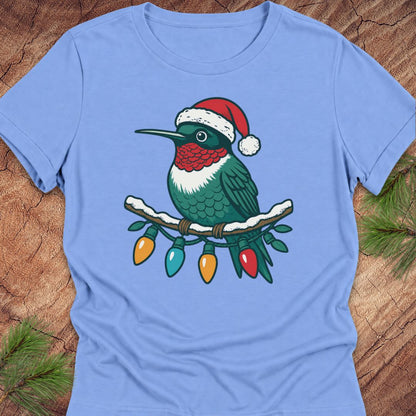 A light blue short-sleeve crewneck t-shirt laid flat on a rustic wood background with pine needles. The shirt features a graphic of a green hummingbird wearing a red and white Santa hat, perched on a branch with hanging Christmas lights.