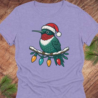 A lavender short-sleeve crewneck t-shirt featuring a graphic of a green hummingbird wearing a red Santa hat and perched on a branch with colorful Christmas lights, displayed flat on a rustic wood background with pine needles.