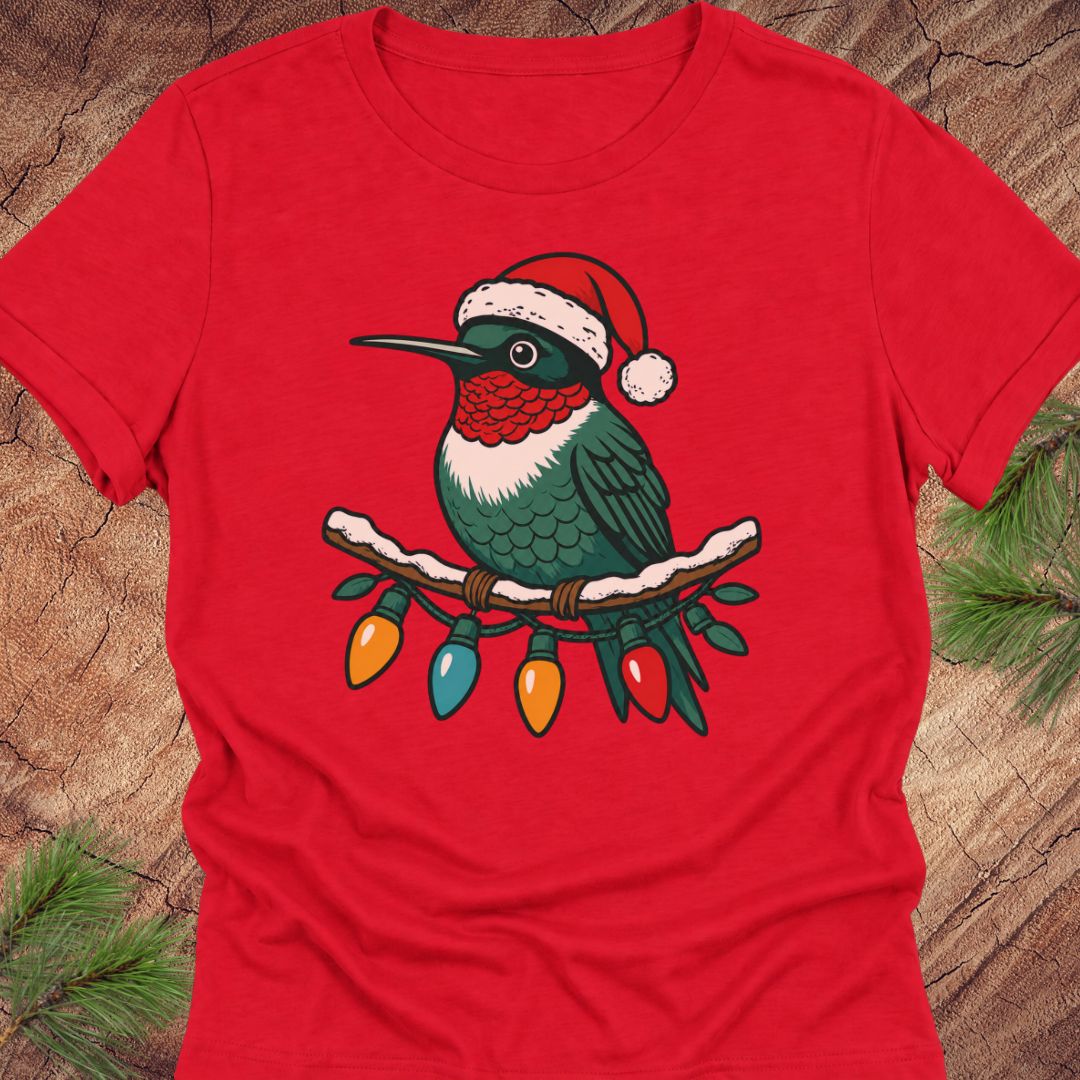 A red short-sleeve crewneck t-shirt featuring a graphic of a hummingbird wearing a Santa hat and perched on a branch with hanging Christmas lights, displayed flat on a rustic wood background with pine needles.