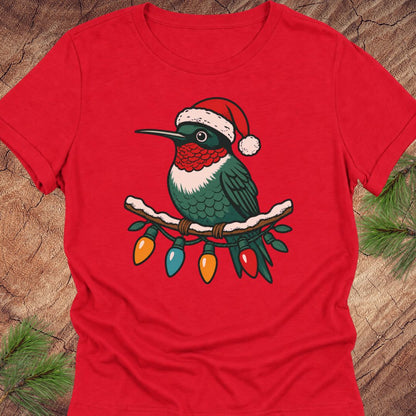 A red short-sleeve crewneck t-shirt featuring a graphic of a hummingbird wearing a Santa hat and perched on a branch with hanging Christmas lights, displayed flat on a rustic wood background with pine needles.