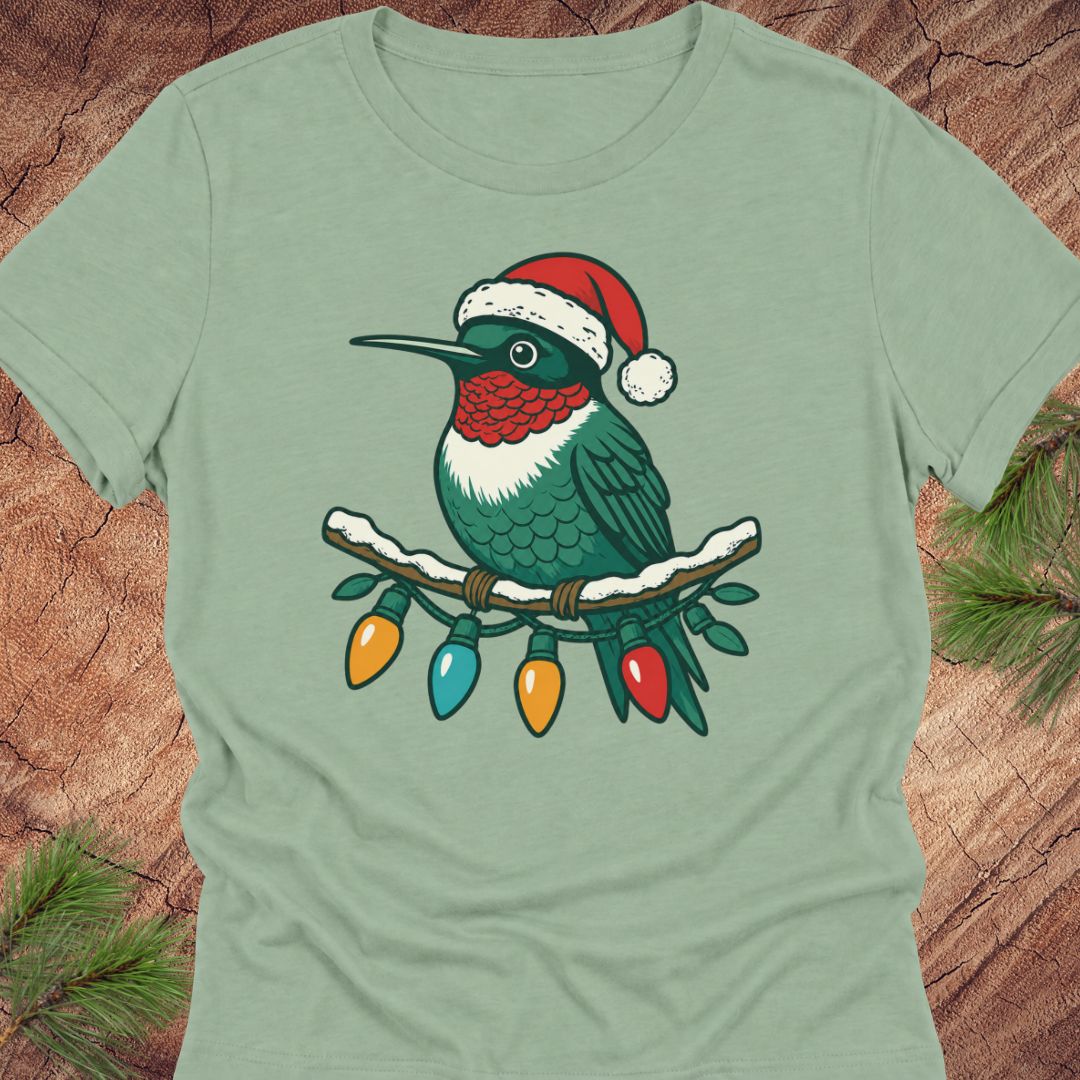 A sage green short-sleeve t-shirt featuring a graphic of a hummingbird wearing a red Santa hat and perched on a branch with colorful Christmas lights, displayed flat on a rustic wood background with pine needles.
