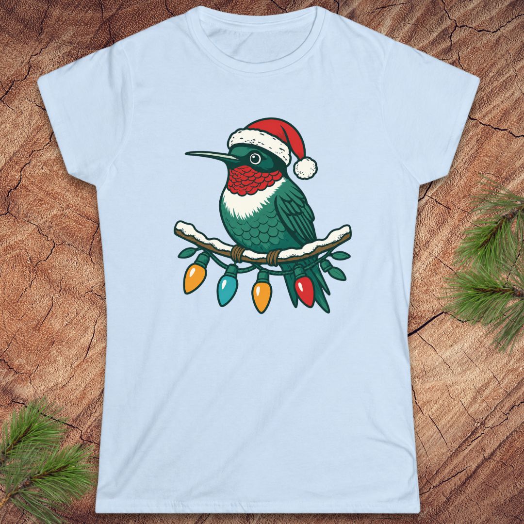 A light blue short-sleeve crewneck t-shirt featuring a graphic of a green hummingbird wearing a red Santa hat and perched on a branch with colorful Christmas lights, laid flat on a rustic wood background with pine needles.