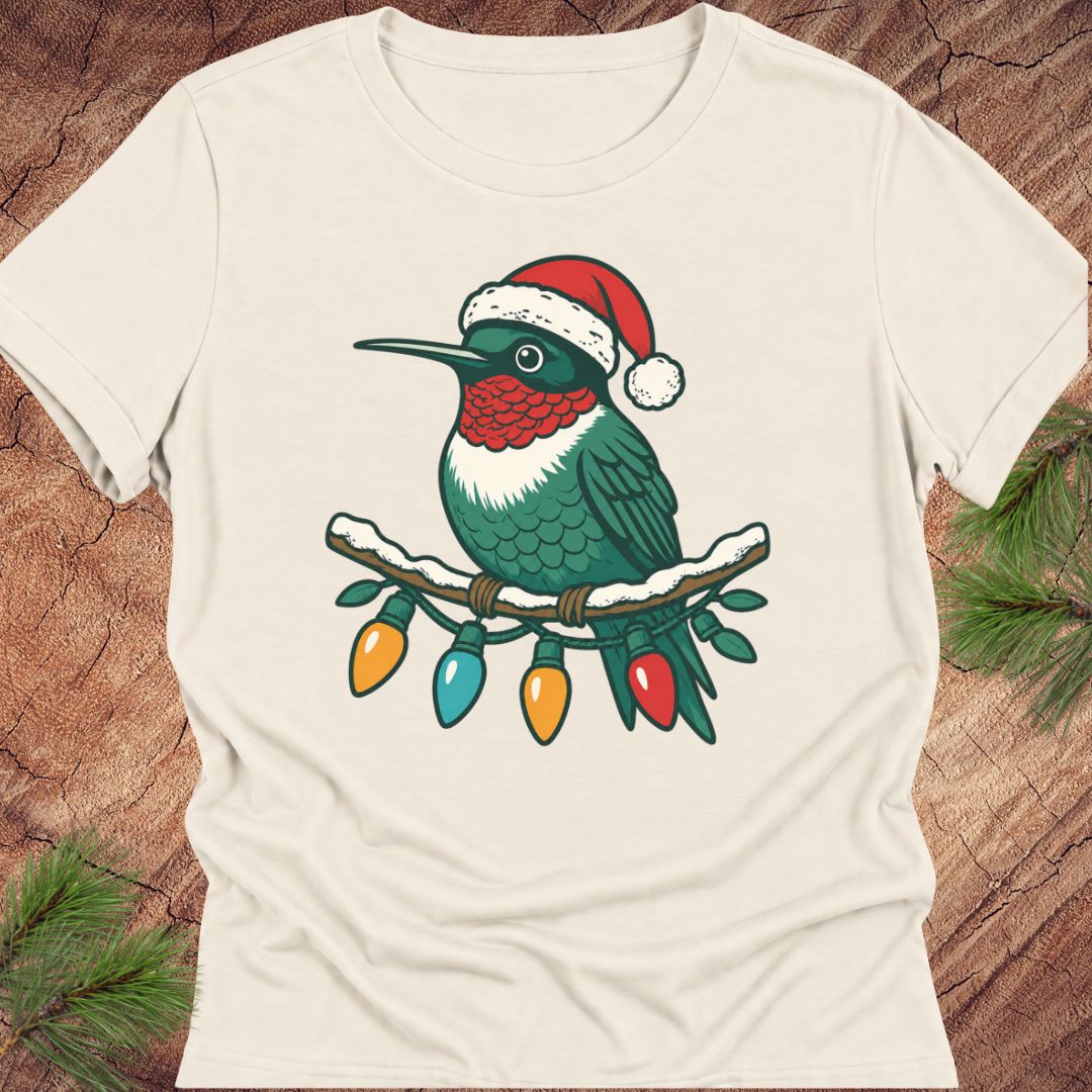 An off-white short-sleeve t-shirt laid flat on a rustic wood background with pine needles in the corners. The shirt features a graphic of a hummingbird wearing a red Santa hat and perched on a branch with colorful Christmas lights.