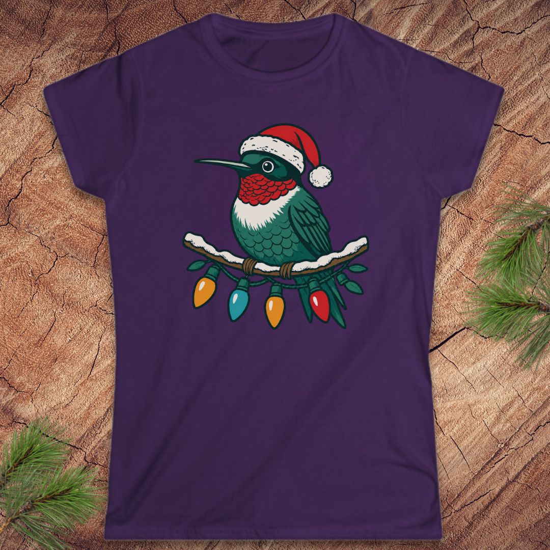 A purple short-sleeve crewneck t-shirt featuring a graphic of a hummingbird wearing a red Santa hat and perched on a branch with Christmas lights, displayed flat on a rustic wood background with pine needles.